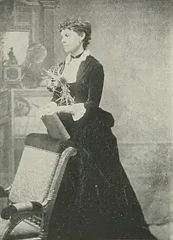 B&W photo of a standing middle-aged woman wearing a dark, high-collared dress with her hair in an up-do.